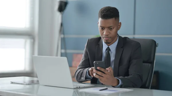Afrikanischer Geschäftsmann nutzt Smartphone während er Laptop im Büro benutzt — Stockfoto