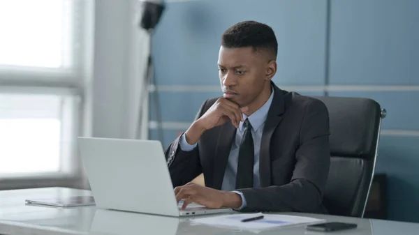 Afrikanischer Geschäftsmann denkt, während er im Büro am Laptop arbeitet — Stockfoto