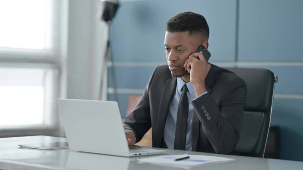 Afrikanischer Geschäftsmann spricht im Büro mit Laptop auf Smartphone — Stockfoto