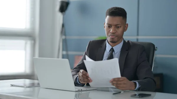 Afrikanischer Geschäftsmann mit Laptop liest Dokumente im Büro — Stockfoto