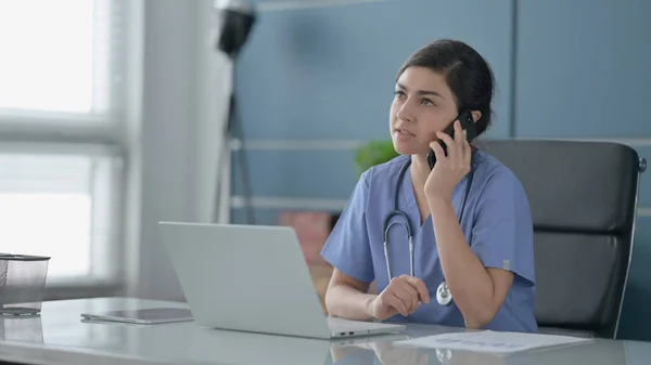 Indiska kvinnliga läkare talar på smartphone när du använder laptop i Office — Stockfoto