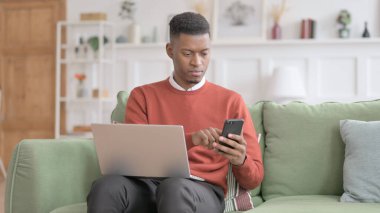 Kanepede akıllı telefon kullanan dizüstü bilgisayarlı Afrikalı adam.