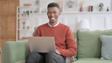 Kanepede dizüstü bilgisayar kullanırken kameraya gülümseyen Afrikalı Adam