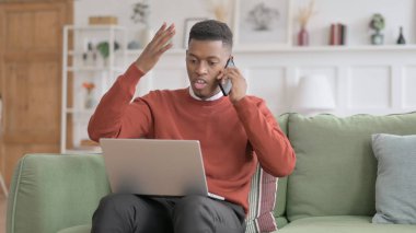Dizüstü bilgisayarlı kızgın Afrikalı adam telefonda konuşuyor.