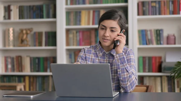 Ofiste dizüstü bilgisayar kullanırken akıllı telefondan konuşan Hintli kadın. — Stok fotoğraf