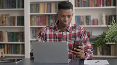 Ofiste dizüstü bilgisayar kullanırken akıllı telefon kullanan Afrikalı adam 