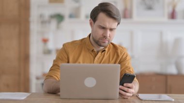 Ofiste dizüstü bilgisayar kullanırken akıllı telefon kullanan genç adam.