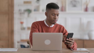 Ofiste dizüstü bilgisayar kullanırken akıllı telefon kullanan Afrikalı adam