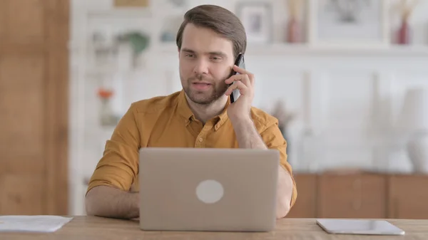 Hombre joven hablando por teléfono mientras usa el ordenador portátil en la oficina —  Fotos de Stock