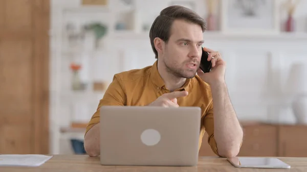 Joven enojado hablando por teléfono mientras usa el ordenador portátil en la oficina — Foto de Stock