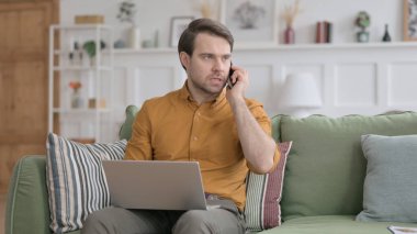 Dizüstü bilgisayarlı kızgın genç adam telefonda konuşuyor.