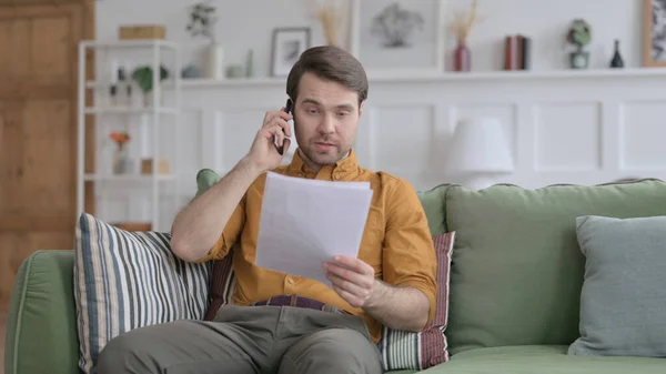 Jeune homme parlant au téléphone tout en lisant des papiers sur le canapé — Photo