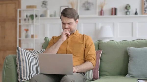 Jeune homme réfléchissant tout en travaillant sur un ordinateur portable sur un canapé — Photo