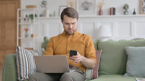 Jeune homme avec ordinateur portable en utilisant Smartphone sur canapé — Photo