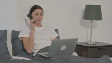Yatakta dizüstü bilgisayar kullanırken telefonda konuşan İspanyol Kadın