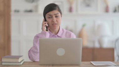 Ofiste dizüstü bilgisayar kullanırken telefonda konuşan kızgın Hintli kadın.