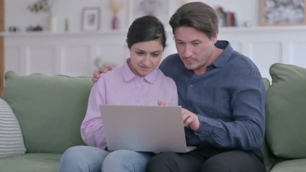 Hombre joven y mujer india usando el ordenador portátil mientras está sentado en el sofá — Vídeo de stock