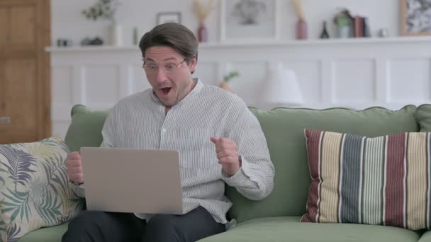 Молодой человек с ноутбуком празднует успех на диване — стоковое видео