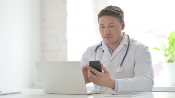 Мужчина врач просмотра Интернета на смартфоне при использовании ноутбука в офисе — стоковое видео