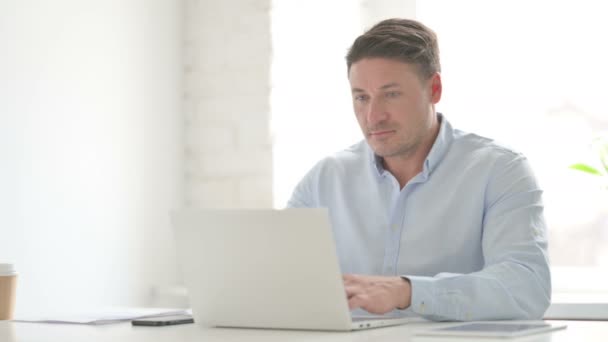 Hombre mirando a la cámara mientras usa el ordenador portátil en la oficina — Vídeos de Stock