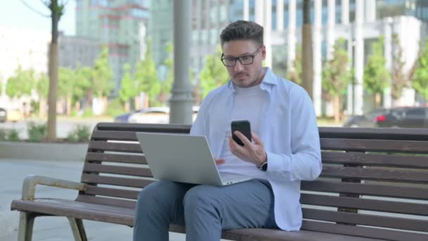 中年人坐在户外长椅上 使用智能手机和笔记本电脑 — 图库视频影像