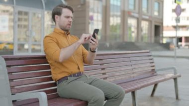 Casual Young Man Browsing Internet on Smartphone while Sitting Outdoor on Bench