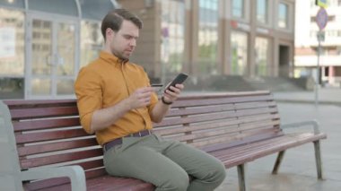 Excited Casual Young Man Shopping Online via Smartphone, Sitting Outdoor