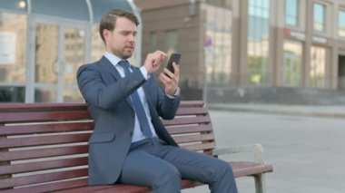 Young Businessman Browsing Internet on Smartphone while Sitting Outdoor on Bench
