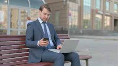Young Businessman Using Smartphone and Laptop while Sitting Outdoor on Bench