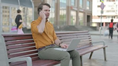 Casual Young Man Talking on Phone and using Laptop while Sitting Outdoor on Bench