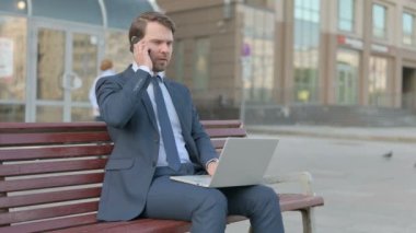 Young Businessman Talking on Phone and using Laptop while Sitting Outdoor on Bench