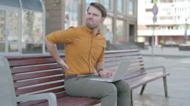 Casual Young Man with Back Pain Using Laptop while Sitting Outdoor on Bench