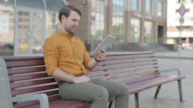 Online Video Chat on Tablet by Casual Young Man Sitting Outdoor on Bench 