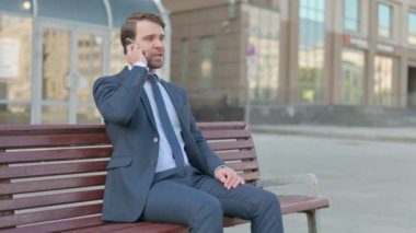 Angry Young Businessman Talking on Phone while Sitting Outdoor on Bench