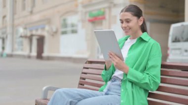Young Woman Celebrating Online Win on Tablet while Sitting Outdoor on Bench