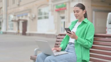 Young Woman Talking on Phone and using Laptop while Sitting Outdoor on Bench