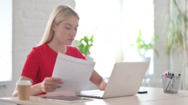 Young Woman Reading Documents and Using Laptop at Work