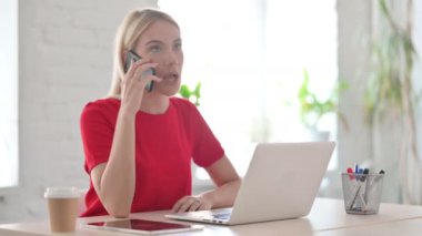Angry Young Woman Talking on Phone while using Laptop in Office