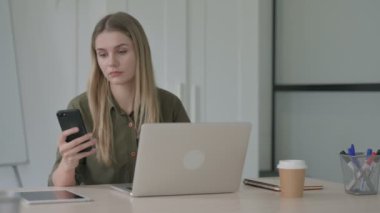 Ofiste dizüstü bilgisayar kullanırken Smartphone 'da internette gezinen Sarışın Genç Kadın 