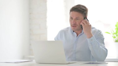 Öfkeli Orta yaşlı adam dizüstü bilgisayar kullanırken telefonla konuşuyor.