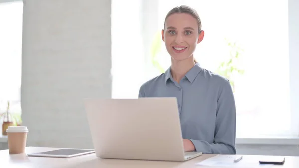 オフィスでノートパソコンを使いながらカメラで微笑む若い女性 — ストック写真