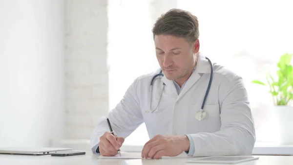 Middle Aged Doctor Thinking while Writing on Paper in Office