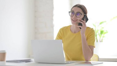 Ofiste dizüstü bilgisayar kullanan genç bir kadın telefonda konuşuyor.