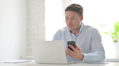 Ofiste dizüstü bilgisayar kullanırken akıllı telefon kullanan orta yaşlı adam