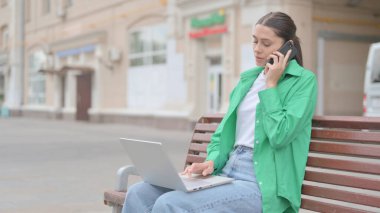 Young Woman Talking on Phone and using Laptop while Sitting Outdoor on Bench