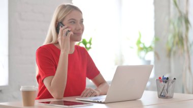 Ofiste dizüstü bilgisayar kullanan genç bir kadın telefonda konuşuyor.