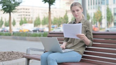 Young Woman Reading Documents and Working on Laptop while Sitting on Bench Outdoor