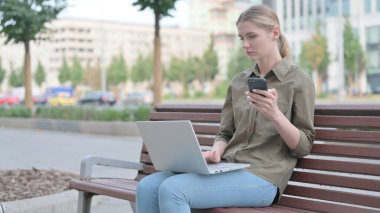Young Woman Using Smartphone and Laptop while Sitting Outdoor on Bench