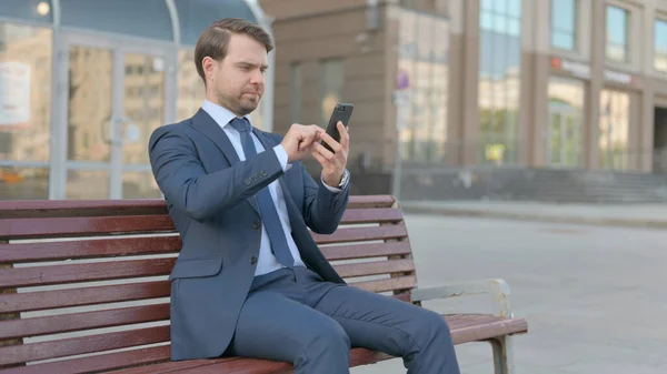 Young Businessman Browsing Internet on Smartphone while Sitting Outdoor on Bench