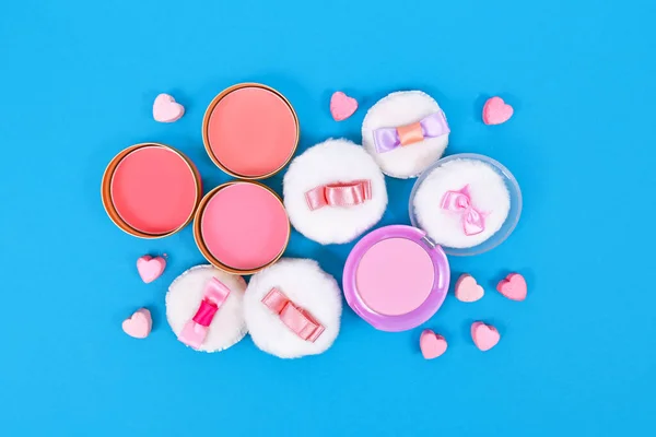 Different pink blush beauty products and powder puffs with ribbons and heart shaped pressed powder on blue background